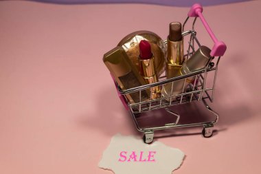 Ruj ve göz farı bir süpermarket arabasının pembe arka planında yatar. El arabasının tekerleklerinin altında küçük bir kağıt parçası var.