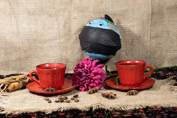 On a jute napkin are two cups of coffee, coffee beans, anise and cinnamon sticks. In the background there is a globe with toy eyes wearing a black medical mask and a purple chrysanthemum.