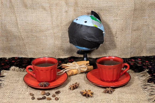 On a jute napkin are two cups of coffee, coffee beans, anise and cinnamon sticks. In the background there is a globe with toy eyes wearing a black medical mask.