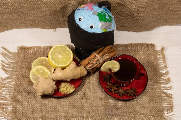There is a cup of tea with lemon, anise and cinnamon sticks on the napkin. Nearby is a saucer of lemon and ginger. In the background there is a globe with toy eyes wearing a black medical mask.