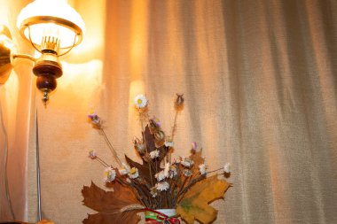 Duvarda bir lamba asılı, altın perdeyi aydınlatıyor. Önümüzde bir buket sonbahar yaprağı ve kurumuş çiçekler var..