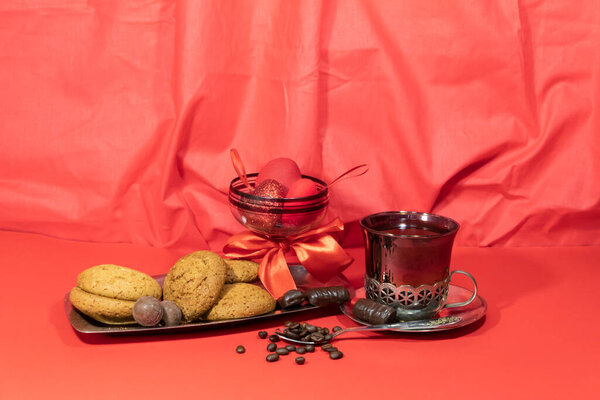 Существует кофе в держатель чашки на красном фоне. Печенье и шоколад на подносе. Неподалеку лежит ложка с кофейными зёрнами. На заднем плане - бокал вина с красным бантом и сердечками.