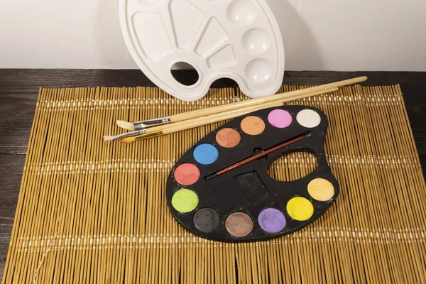 Watercolor paints, palette and paint brushes lie on a bamboo napkin.