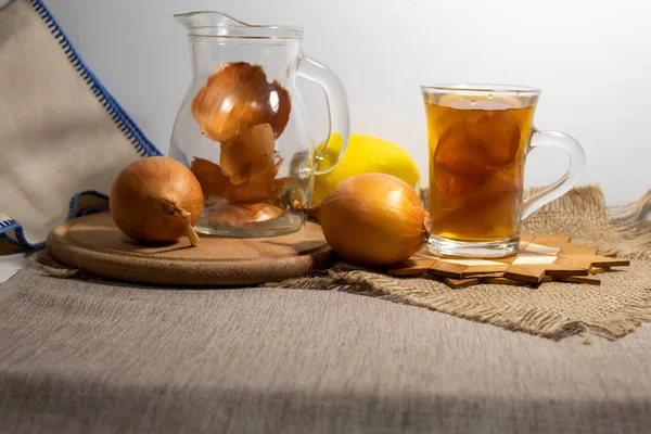 On a jute napkin, on a stand, there is a cup with brewed onion peels and lemon. Nearby is a jug for brewing onion peels and onions and lemon.