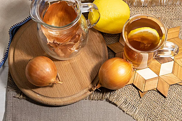 Yukarıdan bak. Yuvarlak bir tahtada soğan, bir limon ve soğan kabuklu bir sürahi bulunur. Yakında bir fincan soğan kabuğu ve limon suyu var.. 