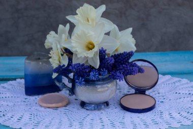 Beyaz bir peçetenin üzerinde bir buket bahar çiçeği, muscari ve nergis var. Yakınlarda mavi bir mum ve pudra var.. 