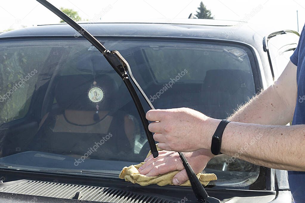 Male hands with a microfiber cloth wipe the windshield of a car, holding the wipers. 
