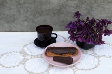 Beyaz bir peçetenin üzerinde bir fincan kahve, bir kek ve bir fincan tabağın üzerinde şeker var. Yakınlarda bir buket çiçek var.. 