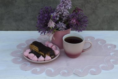 Pembe bir peçetenin üzerinde bir fincan kahve, bir kek ve bir fincan tabağın üzerinde tatlılar var. Yakında pembe bir çiçek kovası var.. 