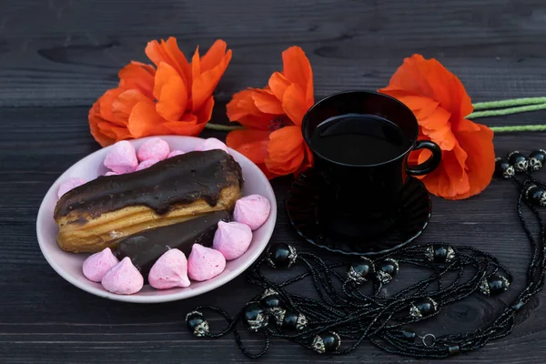 On a dark wooden background, there is a black cup of coffee and a dark necklace. Nearby is a saucer of sweets and cake and three red poppies.