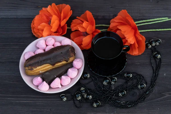View from above. On a dark wooden background, there is a black cup of coffee and a dark necklace. Nearby is a saucer of sweets and cake and three red poppies.