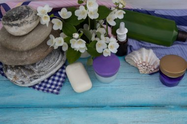 Sabun, krema kavanozları, aromatik yağ, çiçekler ve deniz taşı piramidi mavi bir zeminde uzanır..