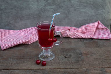 On the table, against the background of a checkered napkin, is a glass with a cherry drink and a straw. Cherries lie nearby.