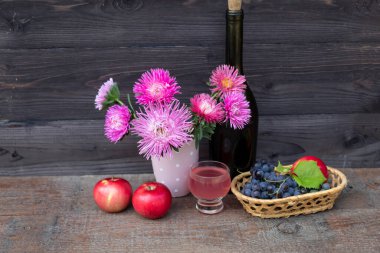 Masanın üzerinde bir buket pembe aster, bir şişe şarap, bir bardak şarap, bir demet üzüm ve elma var..