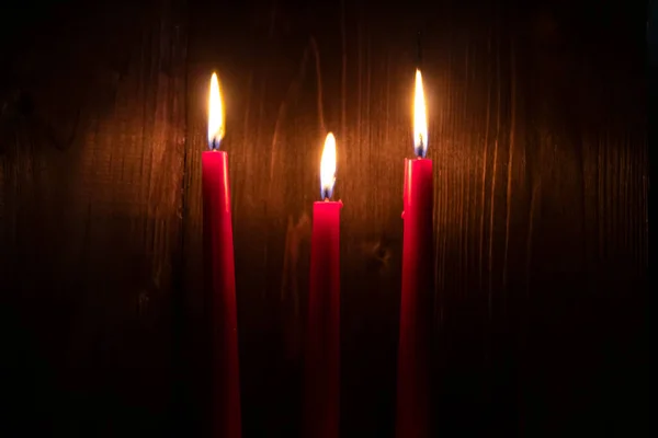 Three red candles are burning in the dark on a dark wooden background.