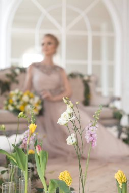 Bahar çiçekleriyle süslenmiş, arka planında bir buket sarı ve beyaz çiçekli bej renkli bir elbise içinde güzel, genç bir gelin. Kapalı alanda zarif bir düğün töreni..