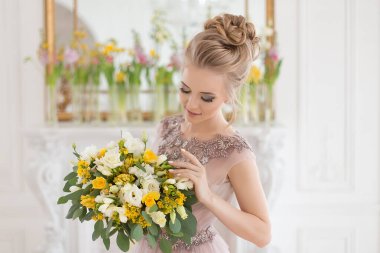 Bahar çiçekleriyle süslenmiş, arka planında bir buket sarı ve beyaz çiçekli bej renkli bir elbise içinde güzel, genç bir gelin. Kapalı alanda zarif bir düğün töreni..