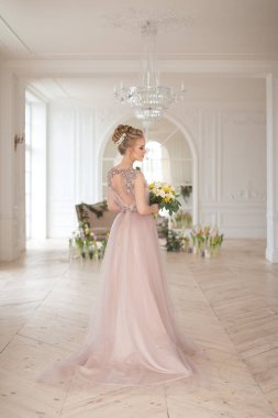 Bahar çiçekleriyle süslenmiş, arka planında bir buket sarı ve beyaz çiçekli bej renkli bir elbise içinde güzel, genç bir gelin. Kapalı alanda zarif bir düğün töreni..