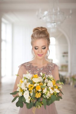 Bahar çiçekleriyle süslenmiş, arka planında bir buket sarı ve beyaz çiçekli bej renkli bir elbise içinde güzel, genç bir gelin. Kapalı alanda zarif bir düğün töreni..