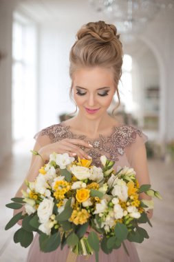 Bahar çiçekleriyle süslenmiş, arka planında bir buket sarı ve beyaz çiçekli bej renkli bir elbise içinde güzel, genç bir gelin. Kapalı alanda zarif bir düğün töreni..