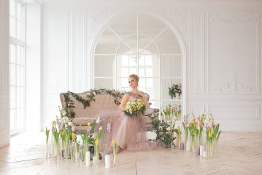 Bahar çiçekleriyle süslenmiş, arka planında bir buket sarı ve beyaz çiçekli bej renkli bir elbise içinde güzel, genç bir gelin. Kapalı alanda zarif bir düğün töreni..