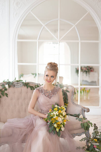 A beautiful young bride in a beige vintage dress with a bouquet of yellow and white flowers on the background of a classic light interior with spring floral decor. Elegant indoor wedding ceremony.