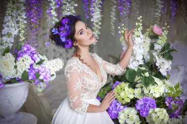 Beyaz dantel bir yatak odası elbisesi, yüksek saç stili mavi şakayıklar, ortanca, mor salkım ve diğer bahar çiçekleriyle süslenmiş klasik bir açık yatak odasında oturan güzel bir genç kız..