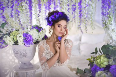 Beyaz dantel bir yatak odası elbisesi, yüksek saç stili mavi şakayıklar, ortanca, mor salkım ve diğer bahar çiçekleriyle süslenmiş klasik bir açık yatak odasında oturan güzel bir genç kız..