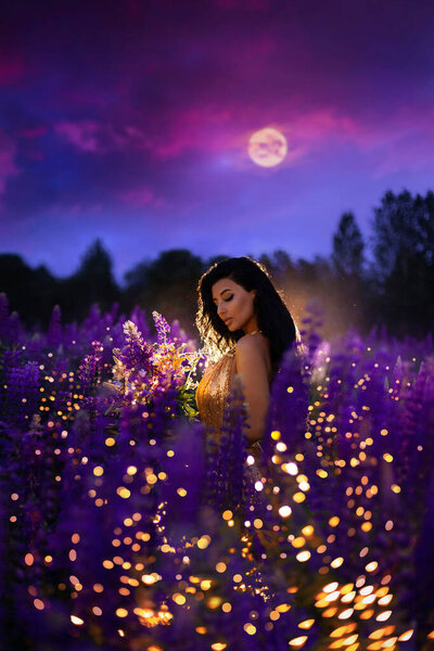 A brunette girl in a golden dress holding a moon in her hands and standing among a blooming purple lupine field with lights and fireflies. A magical night portrait.