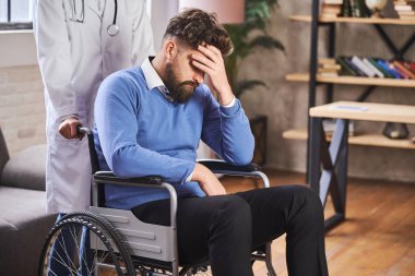 Tekerlekli sandalyede oturan üzgün adam ve onu iten bir doktor.