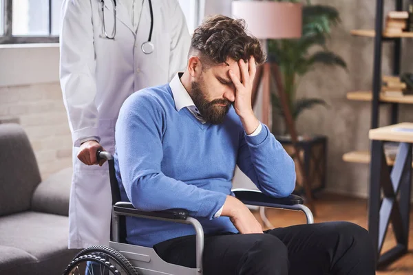 Un uomo triste su una sedia a rotelle che tiene la mano sul viso mentre un medico lo spinge — Foto Stock