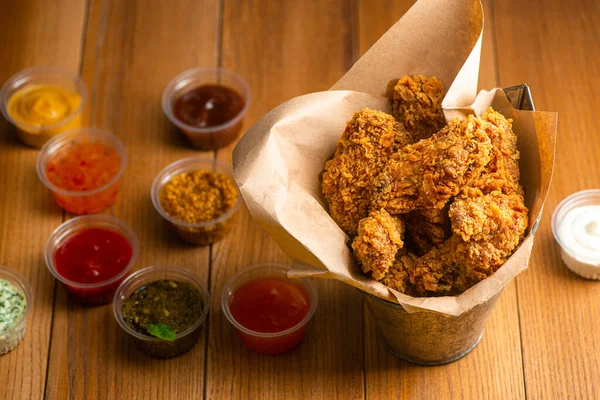 Cubo de alitas de pollo servido con tazas de diferentes salsas en mesa de madera — Foto de Stock