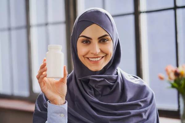 Cheerful Muslim woman holding a bottle of pills and giving a broad smile