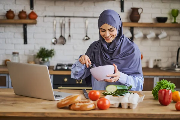 Mujer árabe cocinando una comida y haciendo una videollamada en un portátil —  Fotos de Stock