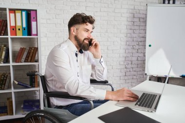 Tekerlekli sandalyede oturan, telefonla konuşan ve dizüstü bilgisayar kullanan neşeli bir adam.
