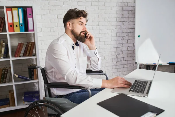 Busy man sitting on a wheelchair, talking on a phone and working on a laptop