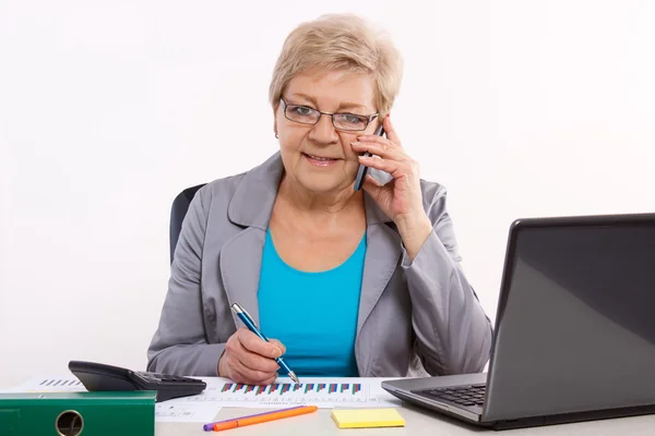 Пожилая деловая женщина разговаривает по мобильному телефону и работает за своим столом в офисе, бизнес-концепция — стоковое фото