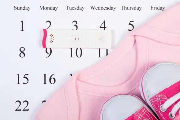 Pregnancy test with positive result and clothing for newborn on calendar, expecting for baby — Stock Photo, Image