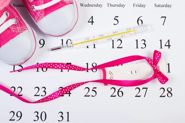 Test di gravidanza con esito positivo, termometro e scarpe da bambino sul calendario, in attesa per il bambino — Foto Stock