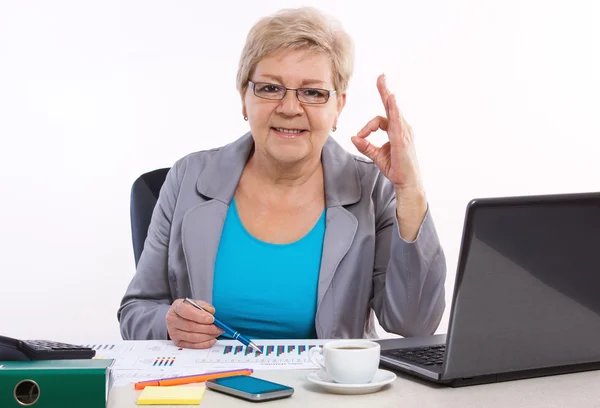Пожилая деловая женщина показывает знак ОК и работает за своим столом в офисе, бизнес-концепции — стоковое фото