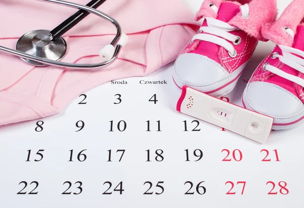 Test di gravidanza con esito positivo, abbigliamento per neonato e stetoscopio sul calendario, attesa per il bambino — Foto Stock
