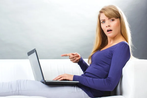 Mujer sorprendida sentada en el sofá y mostrando el ordenador portátil, la tecnología moderna — Foto de Stock