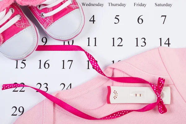 Test di gravidanza con risultato positivo e abbigliamento per neonato sul calendario, in attesa per il bambino — Foto Stock