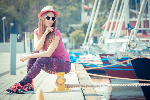 Glad tjej med stråhatt och solglasögon i hamn med båt i bakgrunden, sommartid — Stockfoto