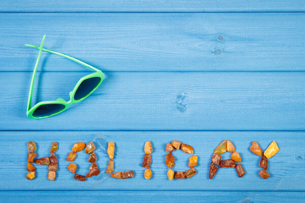 Word holiday made of amber stones and sunglasses on blue boards, summer time, copy space for text