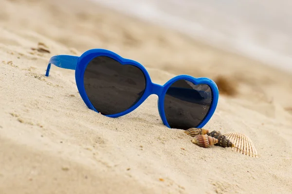 Mavi güneş gözlüğü kum üzerinde kabukları ile kalp şeklinde — Stok fotoğraf