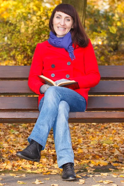 Happy smiling woman sitting on bench reading book in autumn park — Stock Photo, Image