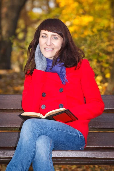 Happy smiling woman sitting on bench reading book in autumn park — Stock Photo, Image