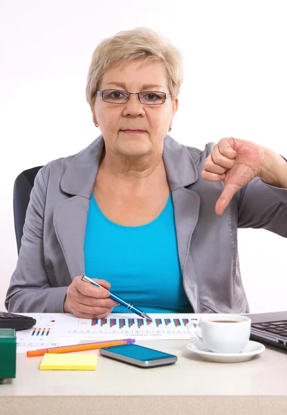Пожилая деловая женщина показывает большие пальцы вниз и работает за своим столом в офисе, бизнес-концепция — стоковое фото