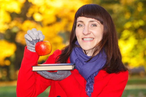 Happy smiling woman holding book and apple in autumn park — Stock Photo, Image